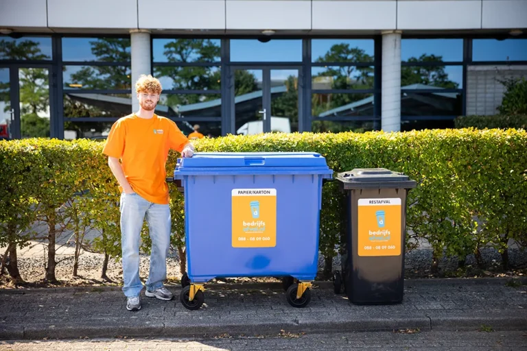 Een bedrijfsafval medewerker staat bij een papier en karton container met daarnaast een restafval container
